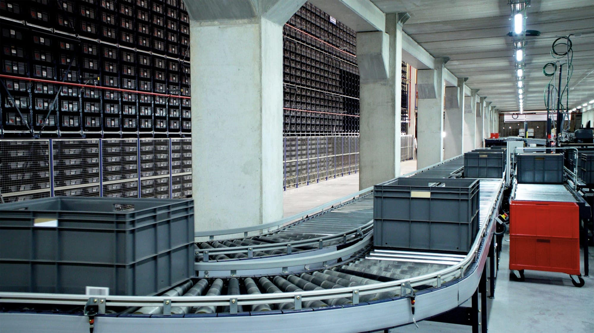 Totes on a curving roller conveyor beside the storage wall of an automated warehouse.